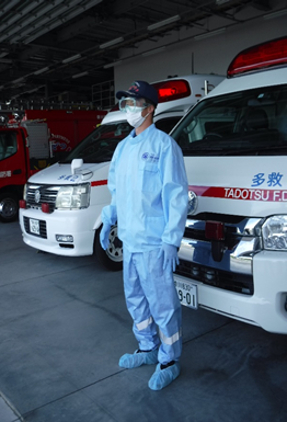 感染症対策用の青色の感染防止着を着用している救急隊員が、救急車の前に立っている写真