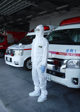 感染症対策用の白色の感染防止着を着用している救急隊員が、救急車の前に立っている写真