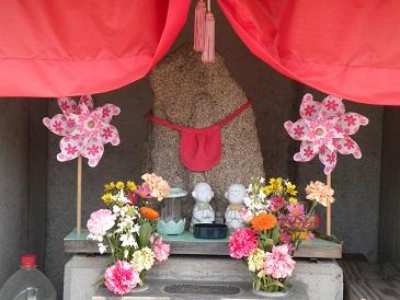赤色のひも付き幔幕の下に供えられた仏花とピンク色の花柄の風車が両端に飾られた後ろに建つ蔵の本地蔵の写真