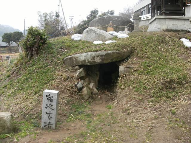 宿地古墳と彫られた石柱の後ろにある細い開口部およびその上に建つ宿地神社の写真