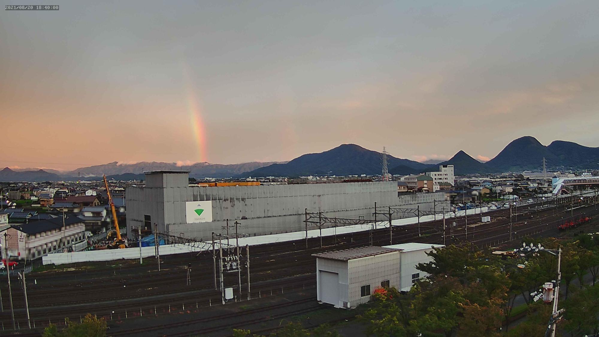 縦に薄く昇っている虹をバックに工事施工会社のロゴが貼られたシートに覆われている多度津町新庁舎を定点カメラで8月20日に撮影した写真